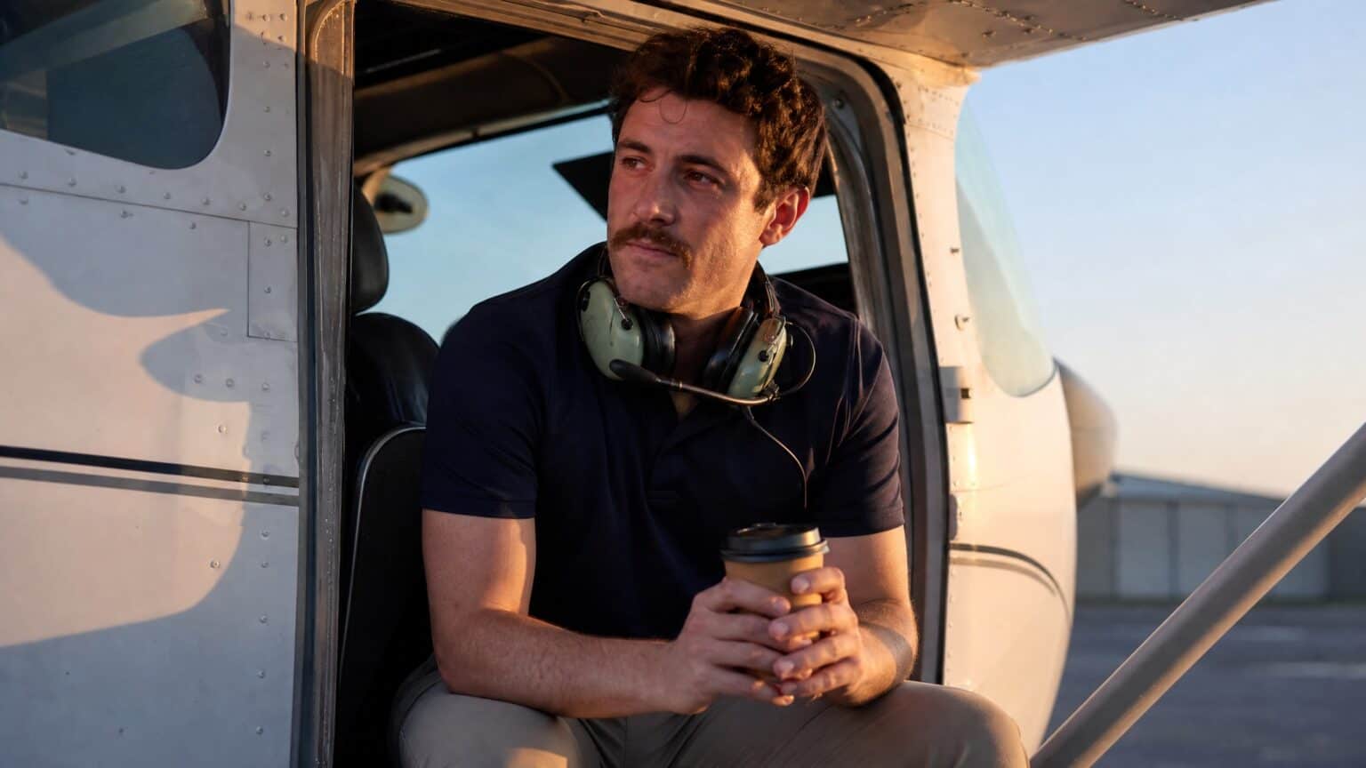 A young flight instructor with a headset around the neck sits in the open door of a small training airplane at sunrise, holding a coffee cup