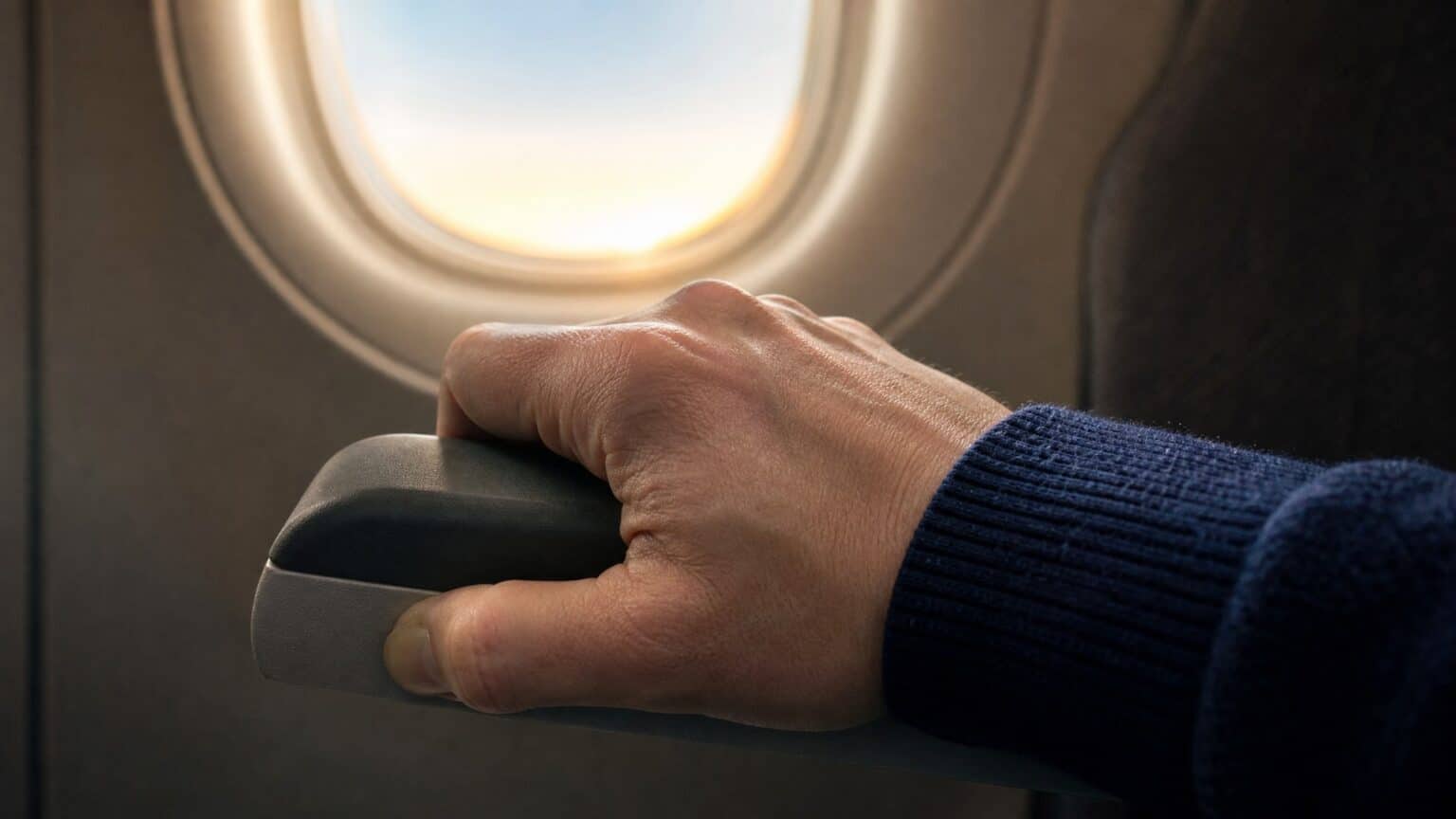 An airline passenger's hand gripping a seat armrest beside a sunlit airplane cabin window during takeoff