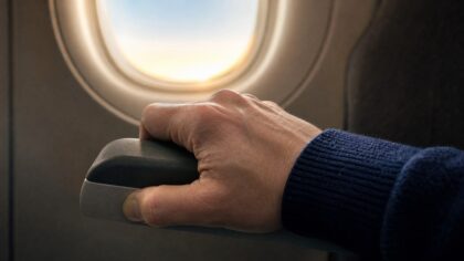 An airline passenger's hand gripping a seat armrest beside a sunlit airplane cabin window during takeoff