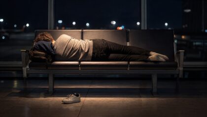 A traveller asleep across a row of airport gate seats at night, shoes off, beneath a pool of warm light