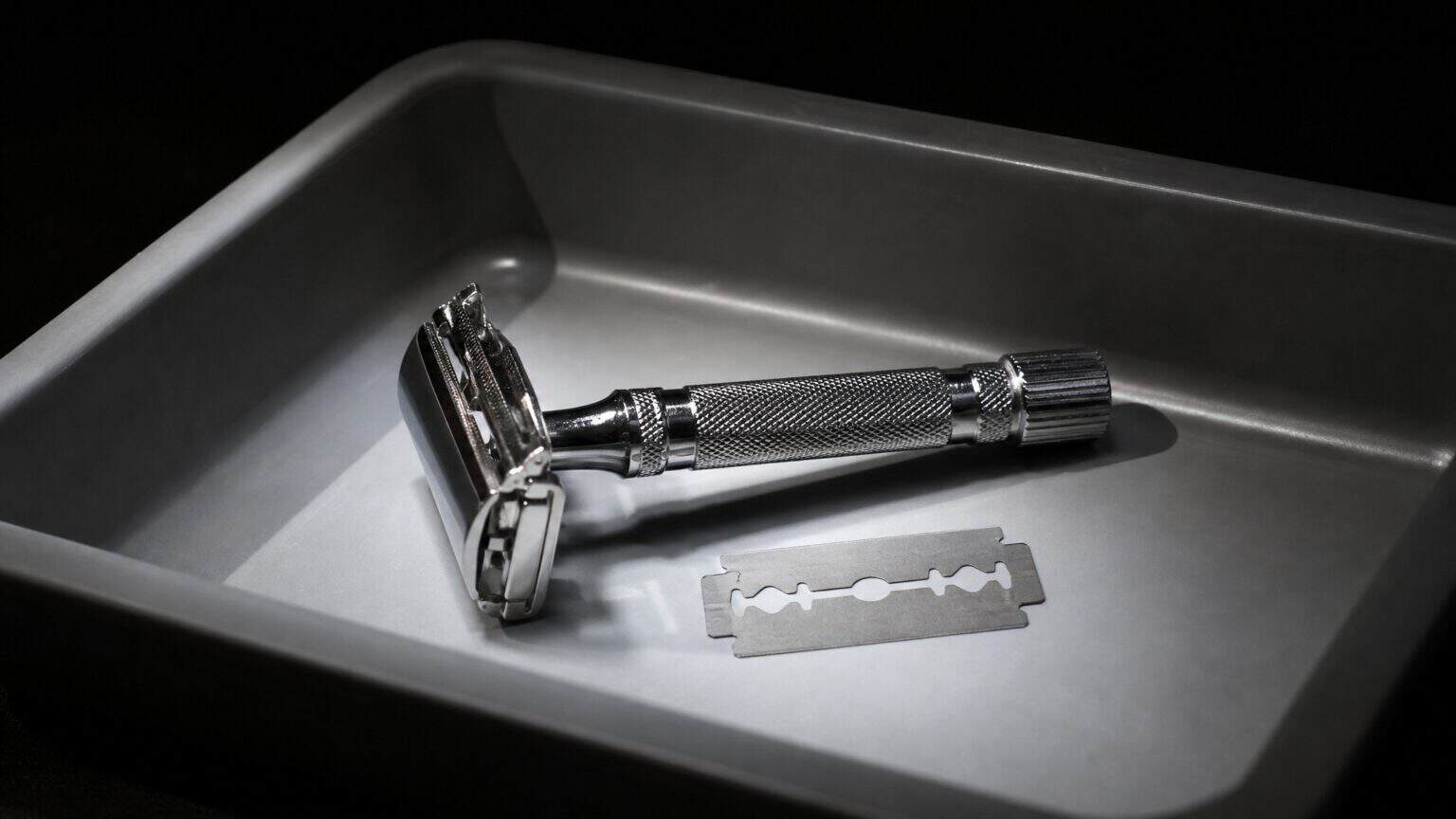 Chrome double-edge safety razor with its loose blade beside it in a grey airport security screening tray