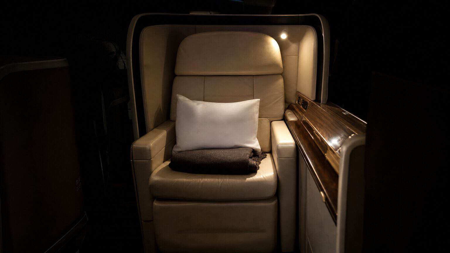 An empty first class airline suite with a cream leather seat, pillow and folded blanket lit by a single reading light in a dark cabin