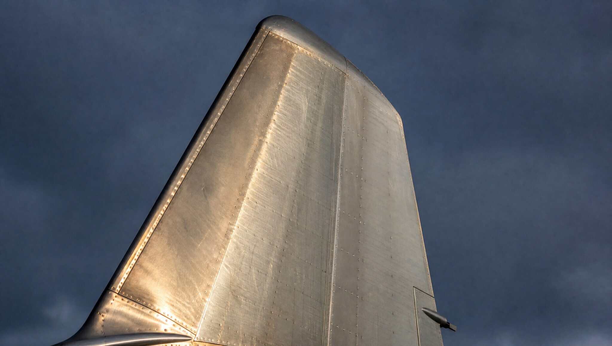 Bare polished aluminium aircraft tail fin with no airline markings, lit by low sun against a dark sky