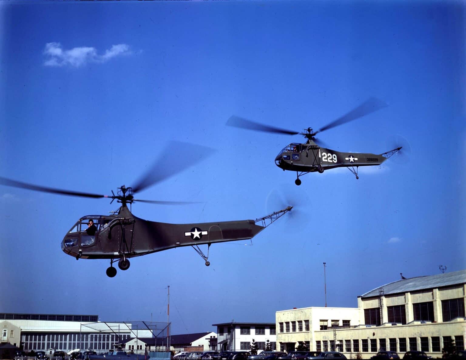 Two US Army Air Forces Sikorsky YR-4B helicopters in flight over a base in about 1944, in color