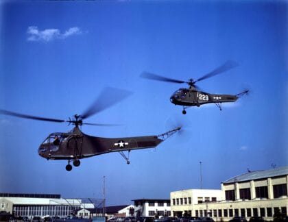 Two US Army Air Forces Sikorsky YR-4B helicopters in flight over a base in about 1944, in color