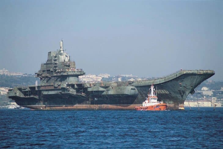 The rusting, unfinished Soviet carrier hull Varyag under tow in Istanbul, with a red tug alongside and no aircraft on deck
