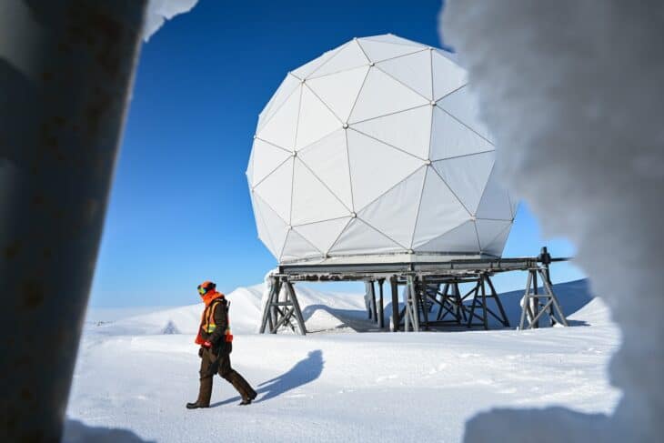 A white geodesic radar dome on a steel platform in the snow, with a bear monitor walking past, at North Warning System site CAM-5A