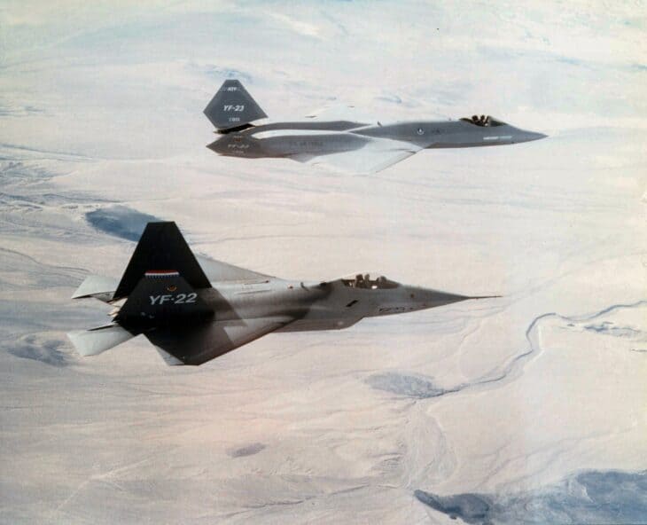 Lockheed YF-22 in the foreground and Northrop YF-23 behind it, flying together over the desert