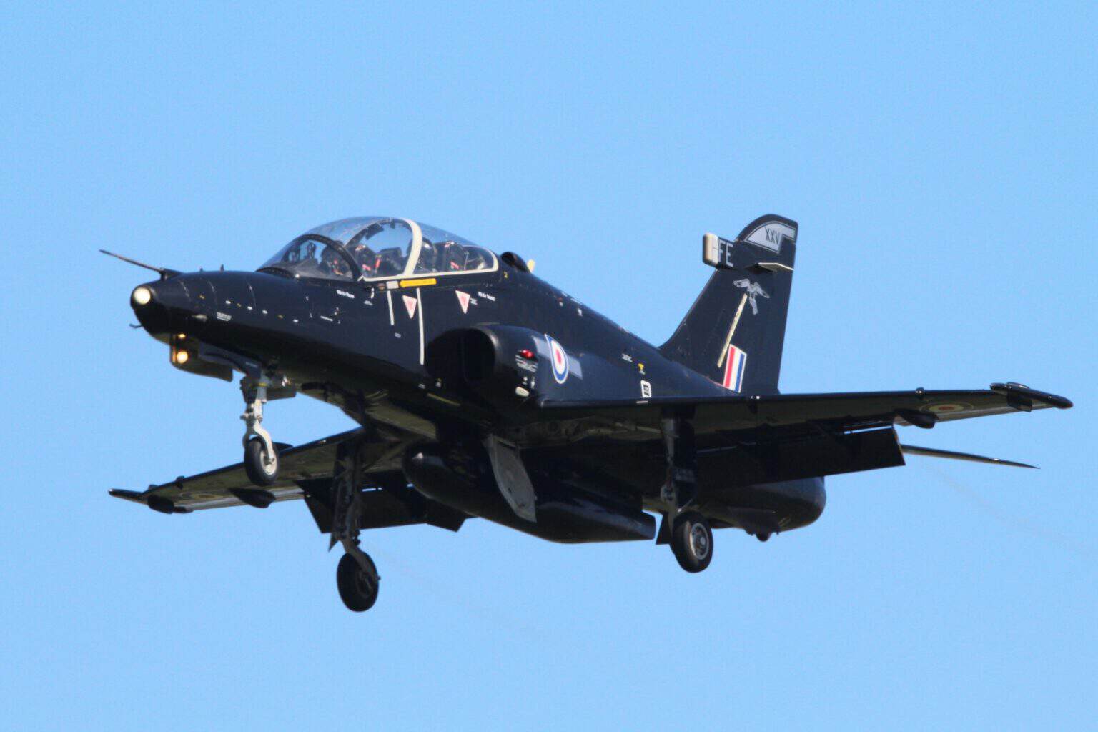 A black RAF Hawk T2 jet trainer flying with its landing gear down against a blue sky at RAF Valley