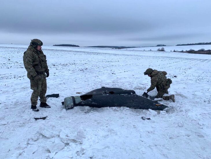 Two Ukrainian police sappers inspect a black delta-wing Russian attack drone lying on a snow-covered field