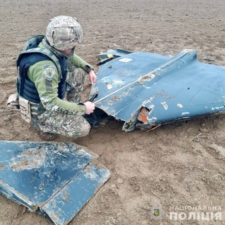 A Ukrainian police officer in a helmet and body armour examines torn blue-grey wing panels from a downed Geran-2 on bare soil