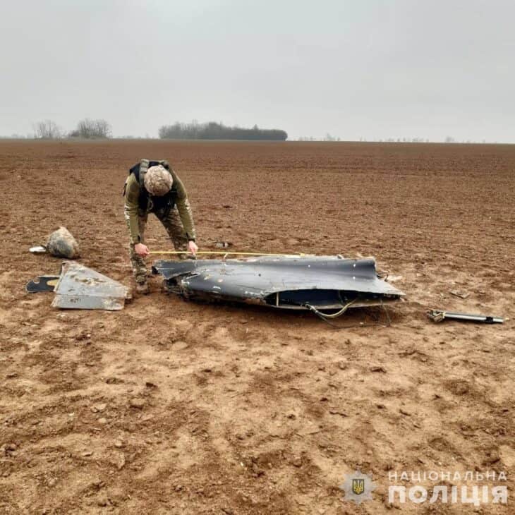 A Ukrainian police officer crouches over the broken dark-grey wing of a downed Geran-2 drone in a ploughed field