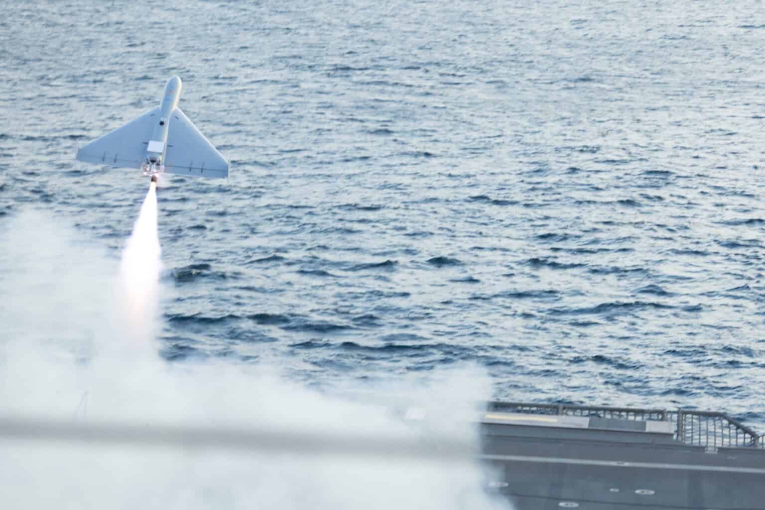 A white LUCAS one-way attack drone climbs away on its rocket booster's plume of smoke above the sea after launching from a US Navy ship