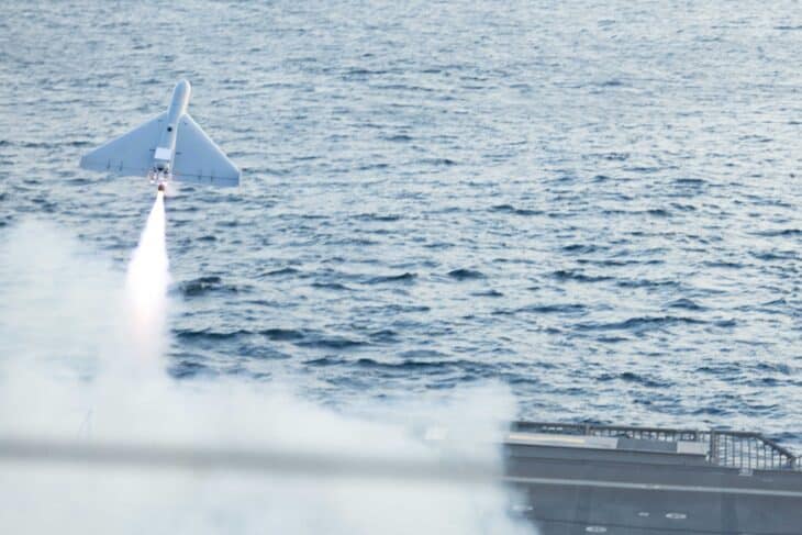 A white LUCAS one-way attack drone climbs away on its rocket booster's plume of smoke above the sea after launching from a US Navy ship