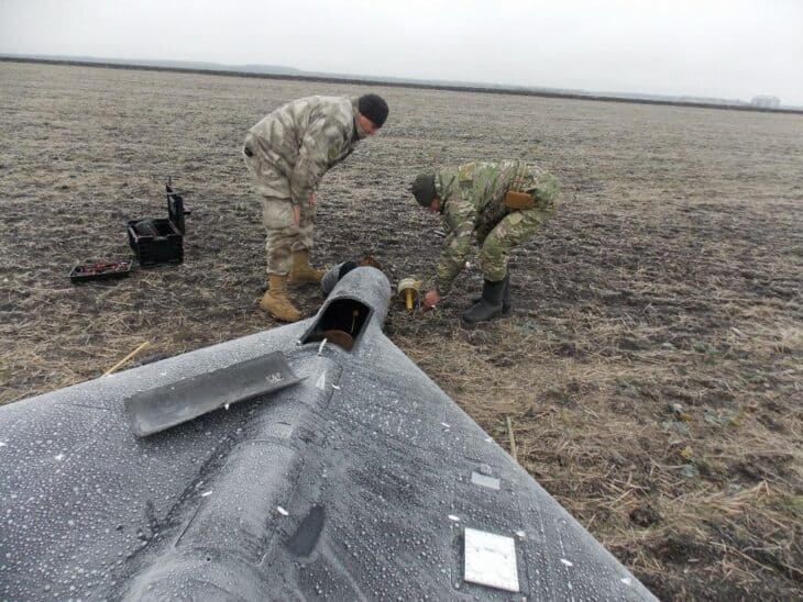 Two Ukrainian police sappers kneel in a field beside the nose and delta wing of a downed Russian attack drone