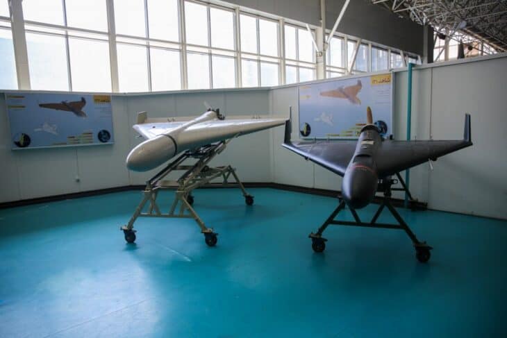 Two delta-wing drones on wheeled stands in an exhibition hall: a larger grey Shahed-136 on the left and a smaller dark Shahed-131 on the right