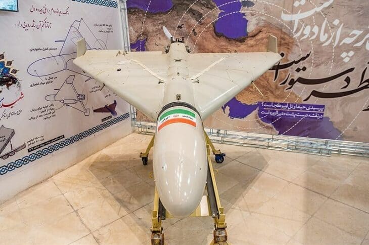 Head-on view of a white Shahed-136 on its stand, showing the pointed nose, cropped delta wing and upturned wingtip fins, in front of a map mural