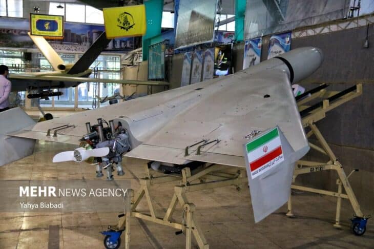 Rear three-quarter view of a grey Shahed-136 on a yellow stand, showing the piston engine and two-bladed pusher propeller at the tail and an Iranian flag on the wing