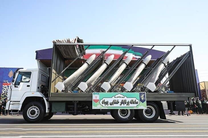 A white truck in a military parade carrying an open launch rack loaded with five delta-wing Shahed drones pointed upward