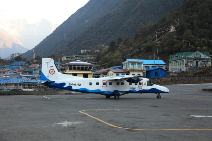 A white and blue Sita Air Dornier 228, registration 9N-AHA, parked on the apron at Lukla's Tenzing-Hillary Airport with the town and mountains behind
