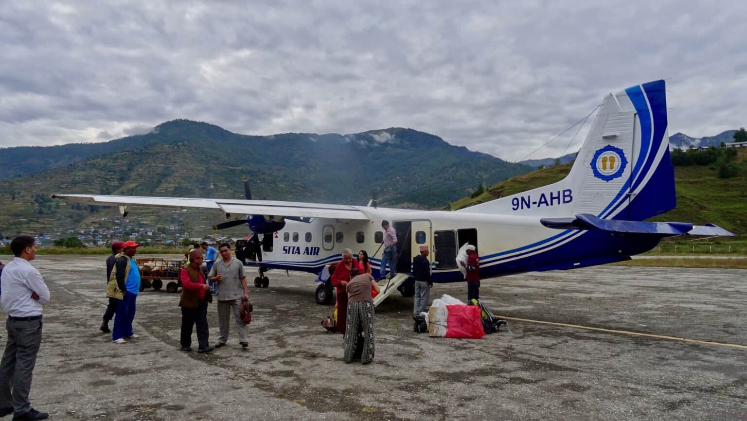 Passengers and baggage beside a white and blue Sita Air Dornier 228, registration 9N-AHB, on the apron at Jumla Airport with forested hills behind