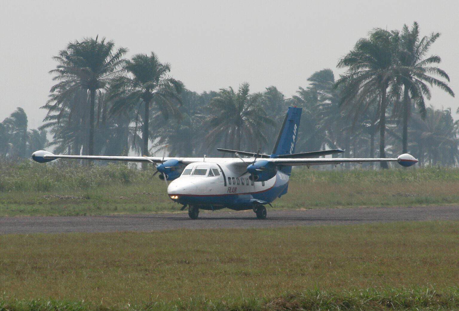 A white and blue Filair Let L-410 Turbolet twin turboprop taxiing on the runway at Kikwit Airport, with palm trees behind