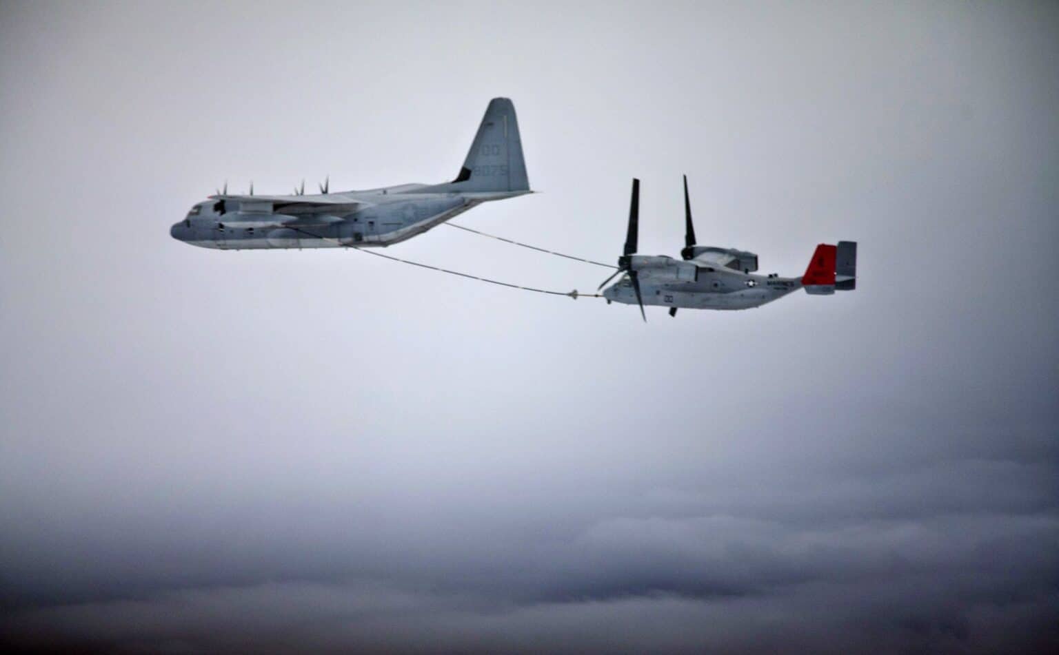 A US Marine Corps KC-130 airplane refueling an MV-22 Osprey tiltrotor in flight above a cloud deck