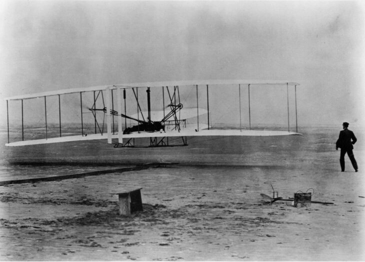 The 1903 Wright Flyer a few feet above the sand at Kitty Hawk with a man standing at the right