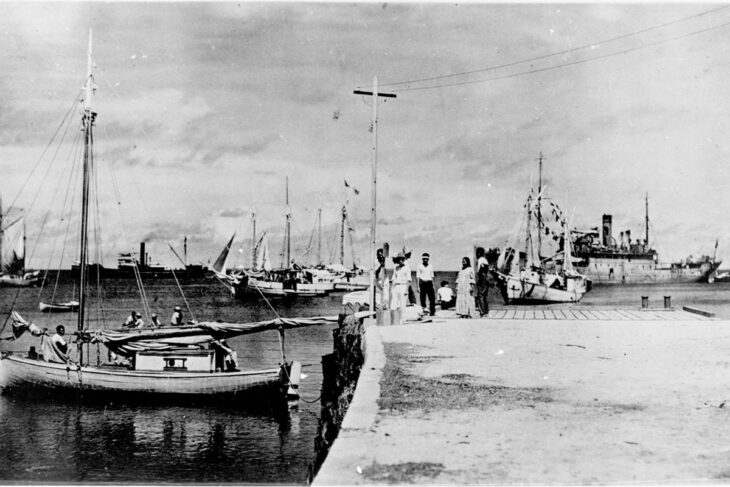 Black and white photo of a dock on Jaluit Atoll with boats moored and a small group of people standing on the pier