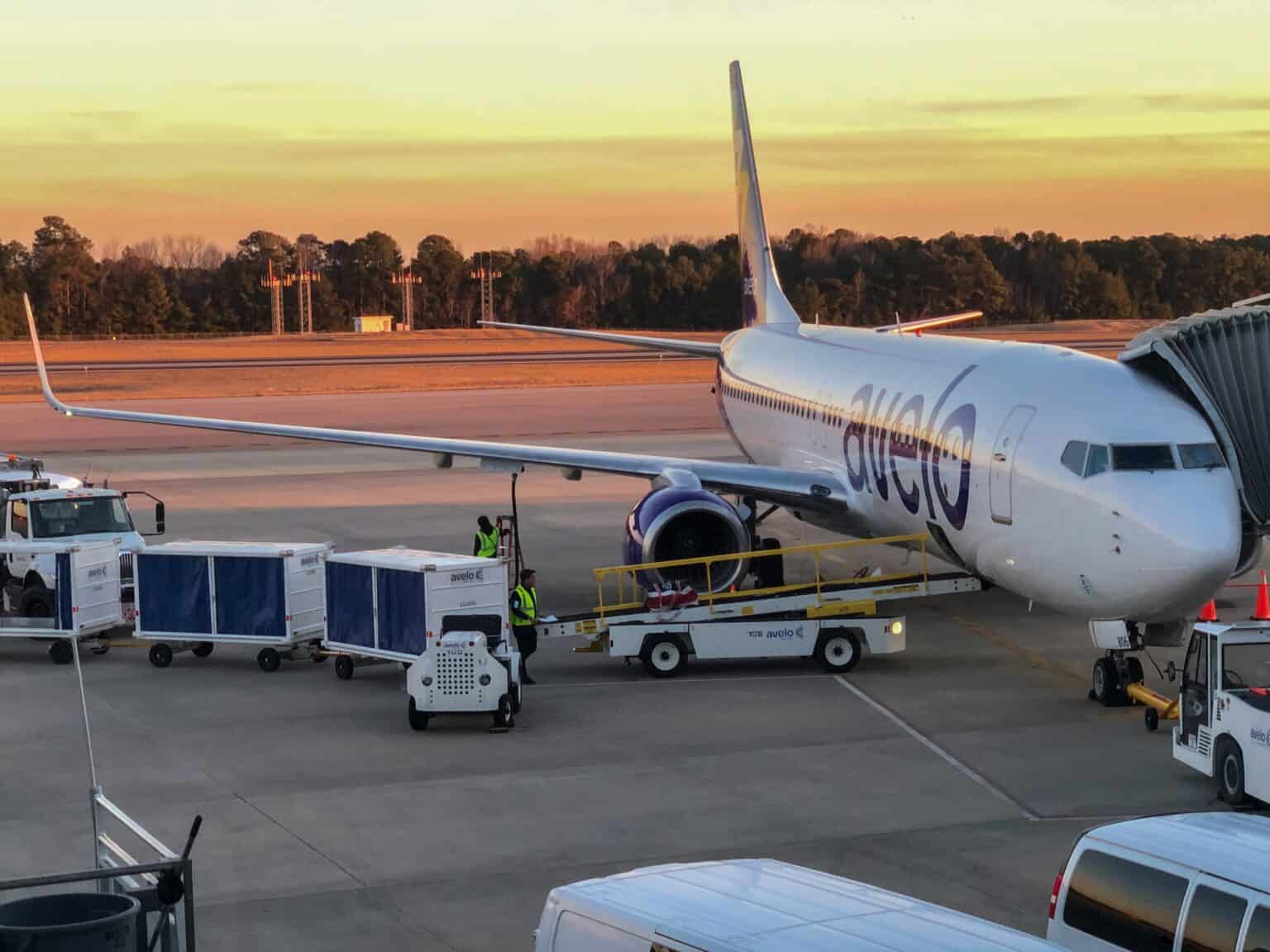 Avelo Airlines Boeing 737-800 N806VL at a jet bridge at Raleigh-Durham International Airport at sunset, with baggage carts and ground crew beside it