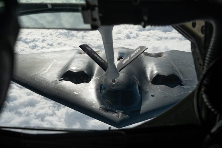 A B-2 Spirit stealth bomber under the refueling boom of a KC-135 tanker, seen from the boom operator's window above a layer of cloud