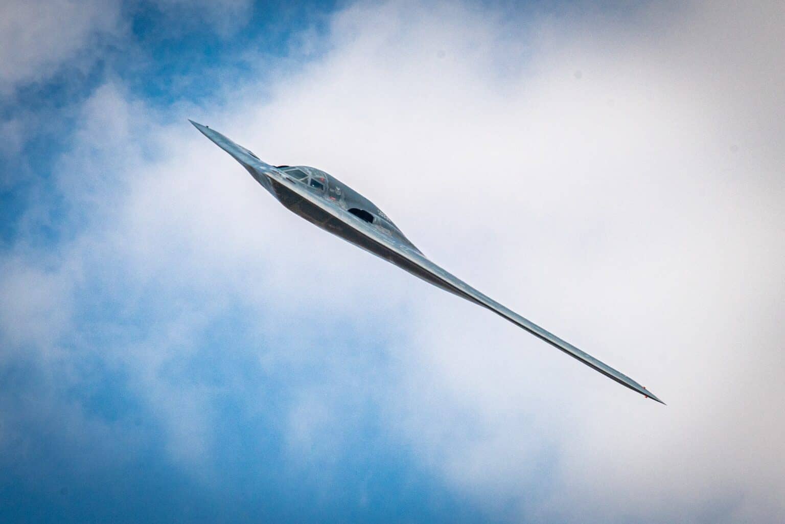 A Northrop Grumman B-2 Spirit stealth bomber banking steeply overhead against a cloudy blue sky