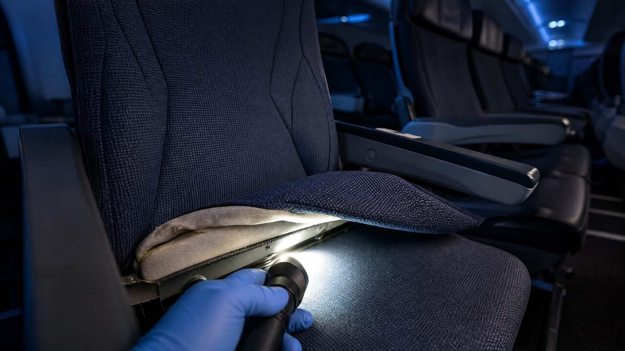 Gloved hand shining a flashlight under a lifted economy seat cushion in a dark airliner cabin