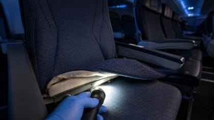 Gloved hand shining a flashlight under a lifted economy seat cushion in a dark airliner cabin
