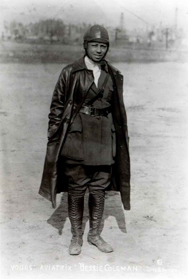 Bessie Coleman in flying cap, jacket and boots, standing in a field in about 1922