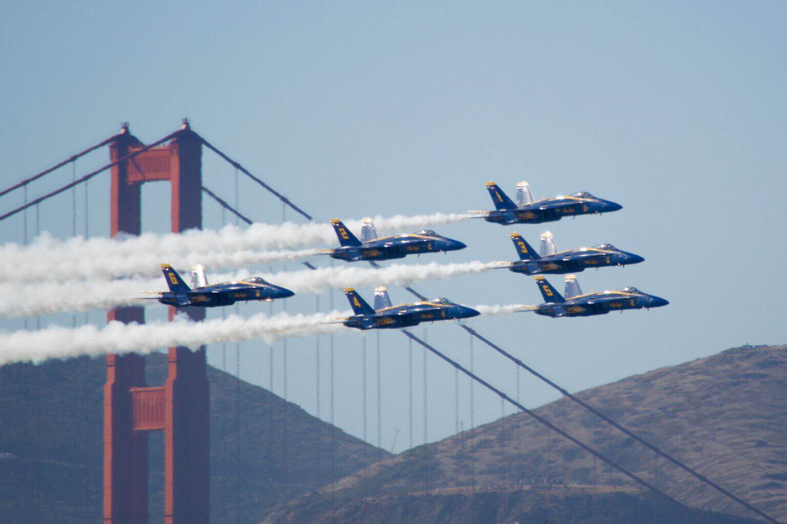 Six US Navy Blue Angels F/A-18 Hornets trailing smoke in formation past a tower of the Golden Gate Bridge