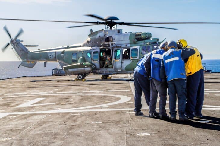 Brazilian Air Force H-36 Caracal helicopter on a ship deck with deck crew in blue and yellow vests