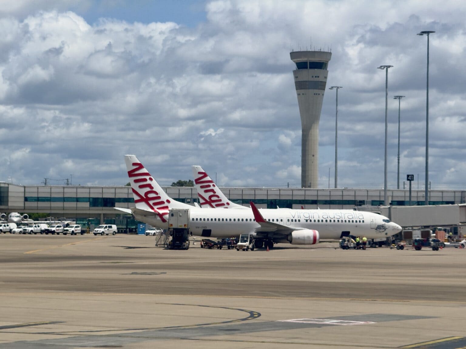 Two Virgin Australia Boeing 737s on the apron at Brisbane Airport's domestic terminal beside the control tower under a cloudy sky