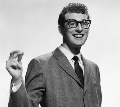 Buddy Holly smiling in his black-framed glasses, suit and tie in a 1950s Brunswick Records publicity portrait
