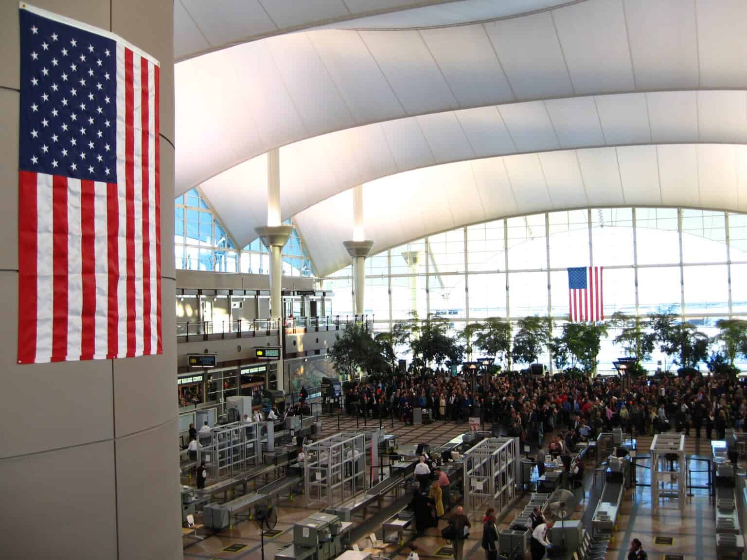 Crowd of passengers queuing at the TSA security checkpoint in the Great Hall of Denver International Airport, with an American flag hanging in the foreground