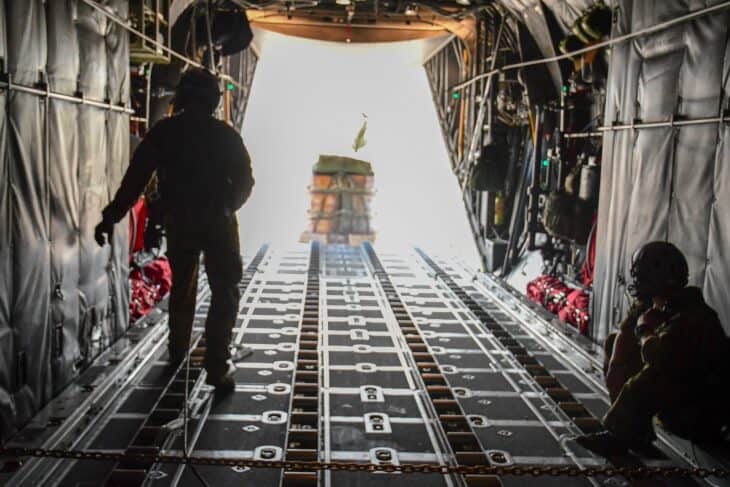 Two loadmasters watch a pallet of cargo slide out of the open rear ramp of a C-130H3 Hercules in flight