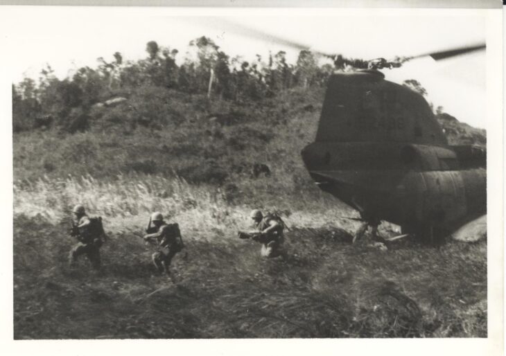 Black and white photo of Marines running through tall grass from a CH-46A Sea Knight that has just landed in Vietnam