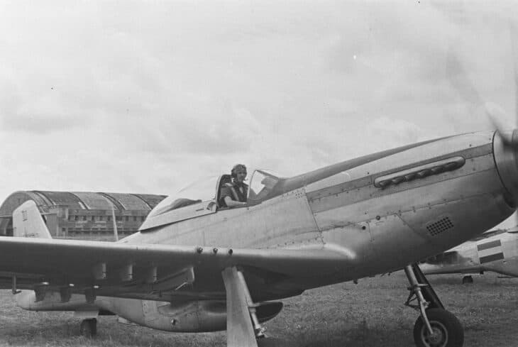 A North American P-51 Mustang fighter in wartime black and white, parked with its pilot in the cockpit