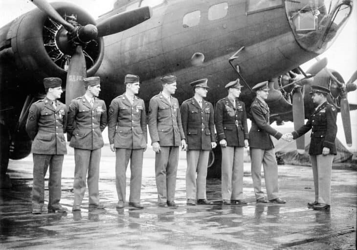305th Bombardment Group officers lined up on a wet English ramp beneath a B-17, shaking hands with a visiting officer