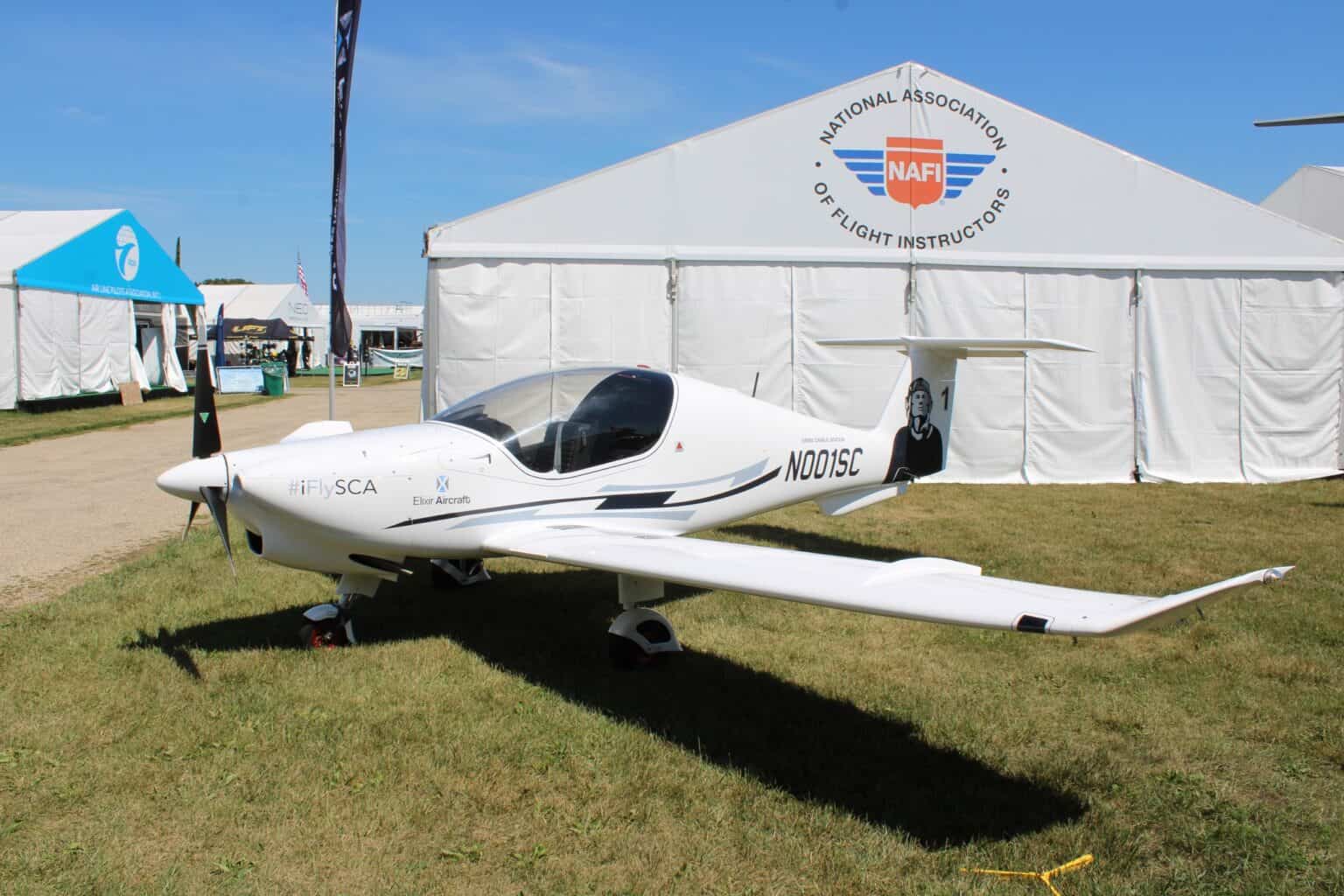 White Elixir Aircraft Elixir two-seat trainer N001SC parked on grass in front of a display tent