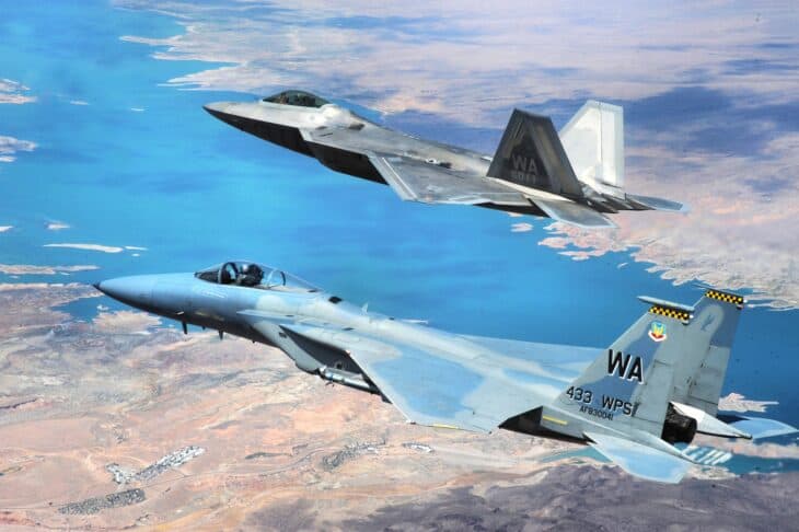 A Lockheed Martin F-22 Raptor flying above an F-15 Eagle of the 433rd Weapons Squadron over a lake and desert