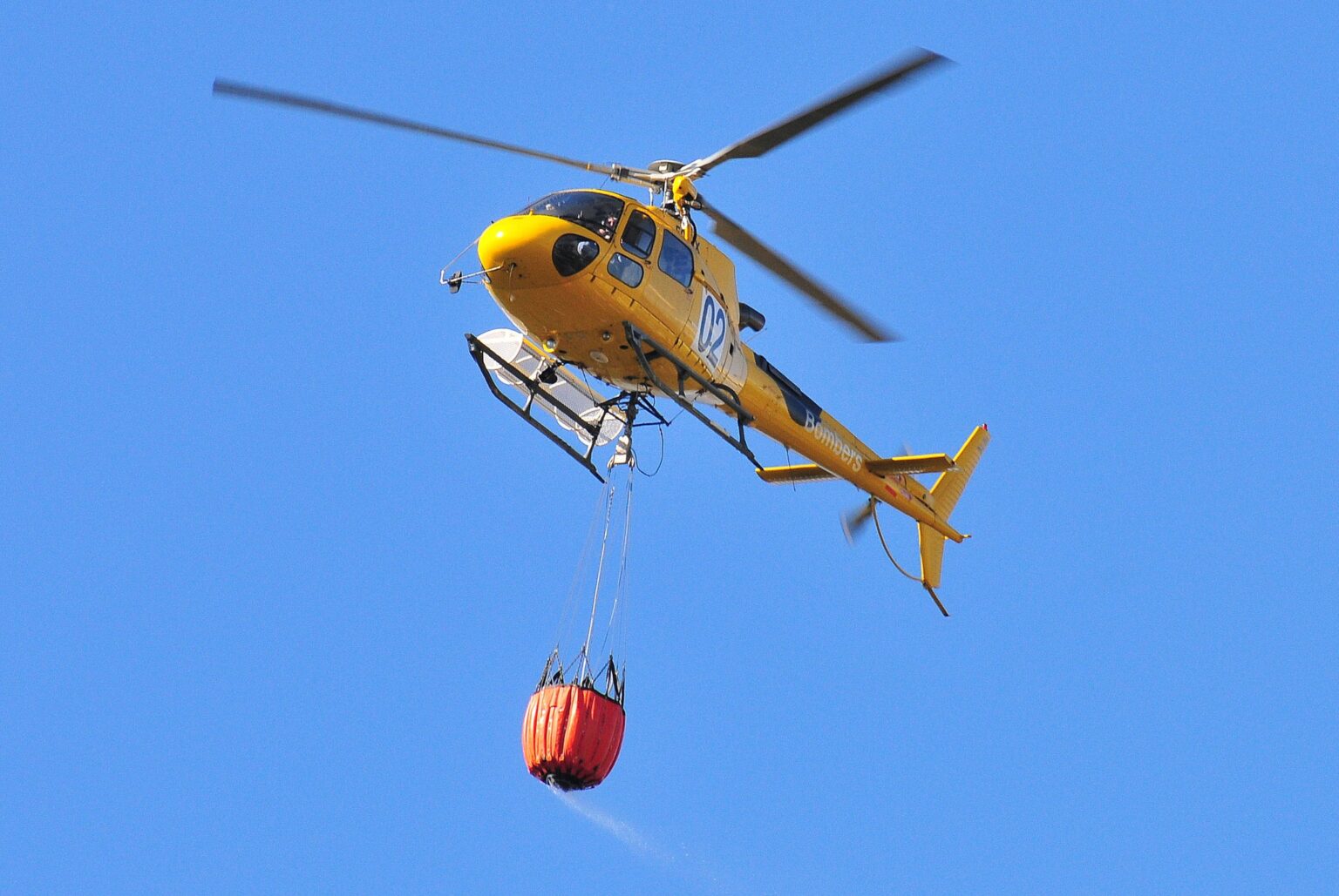 Yellow Airbus AS350 B3 Ecureuil helicopter in flight with an orange water bucket slung beneath it