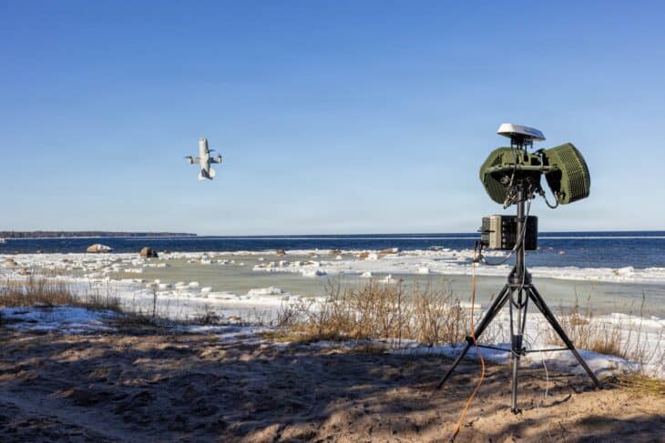 A US Army counter-drone defense system on a tripod beside a shoreline as a drone flies overhead during a test