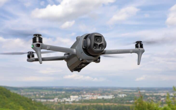DJI Mavic 4 Pro in flight with its triple-lens camera pod facing the camera over a hilly landscape