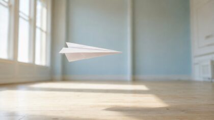 A white paper airplane gliding level through a bright high-ceilinged room in warm morning light